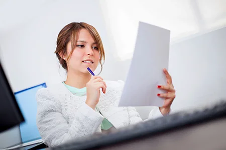 A woman at a desk reads a printed document with a pen held to her lips