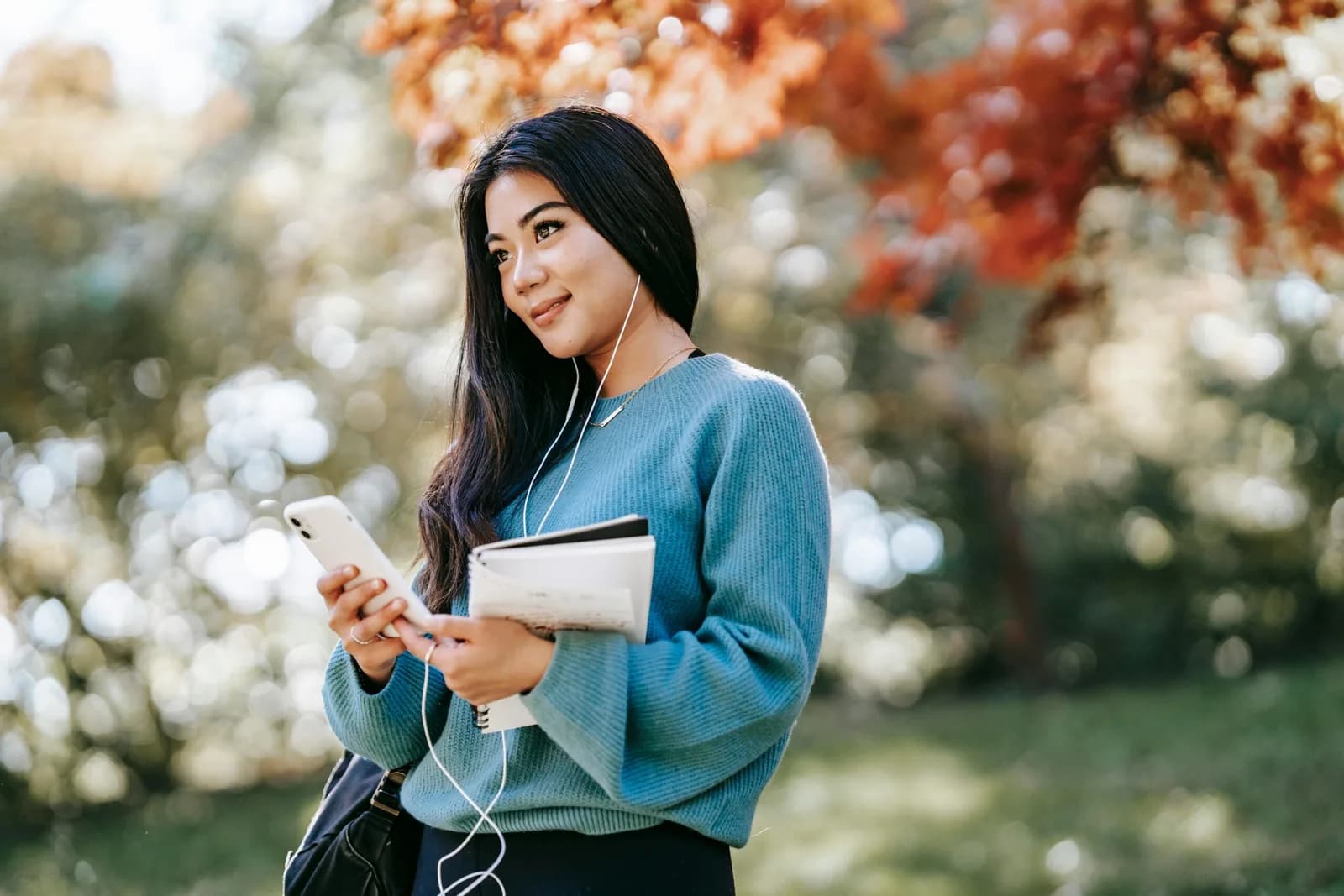 A student in a blue sweater holds a phone and notebook outdoors among autumn trees