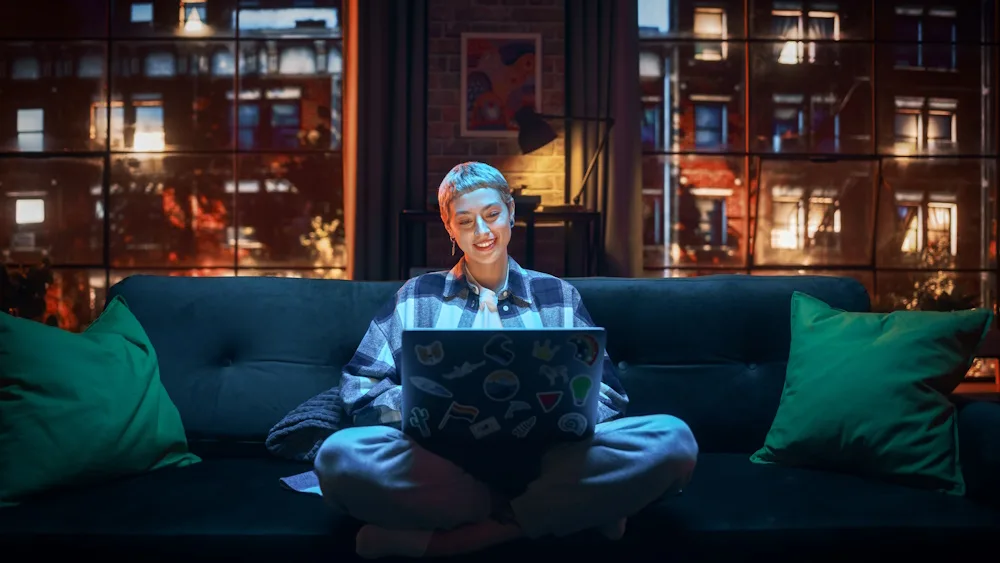 A person sits cross-legged on a couch with a sticker-covered laptop, city windows behind