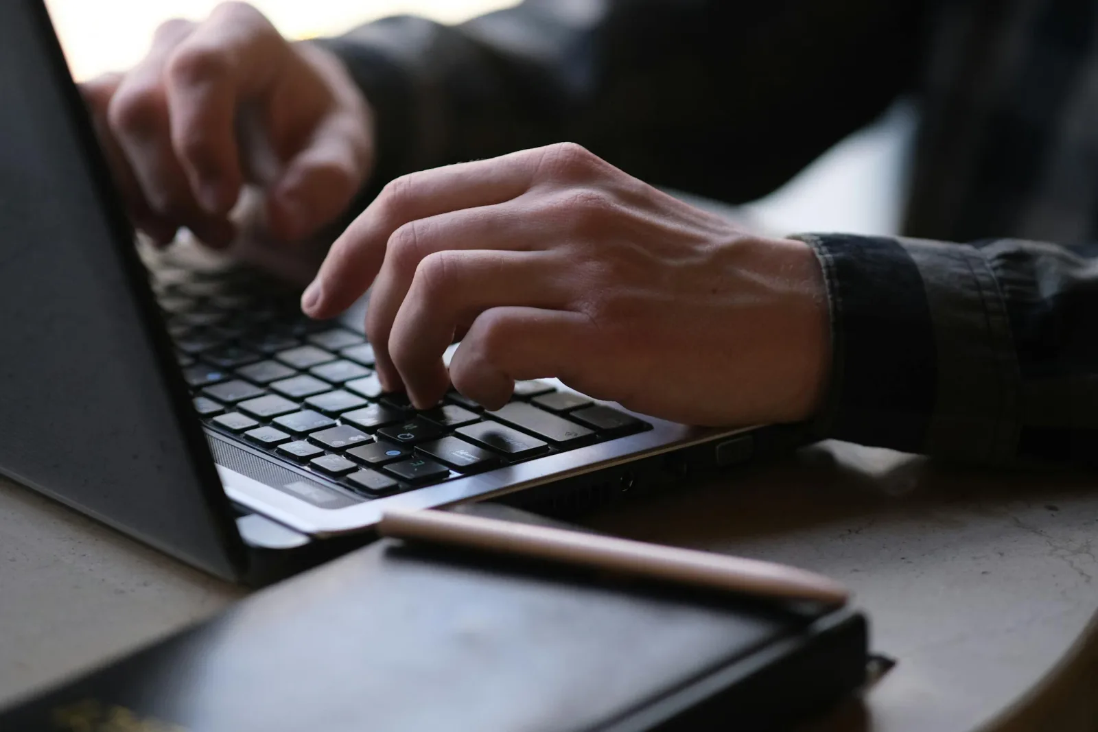 A close-up of two hands typing on a laptop keyboard beside a notebook and pen