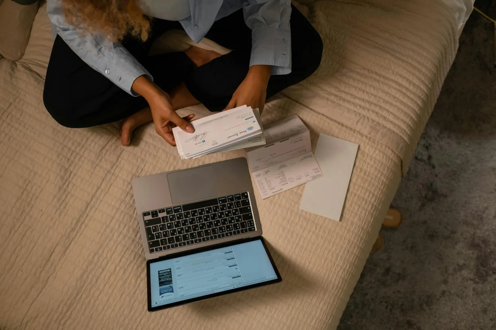An overhead view of a person sorting paper bills beside an open laptop on a bed