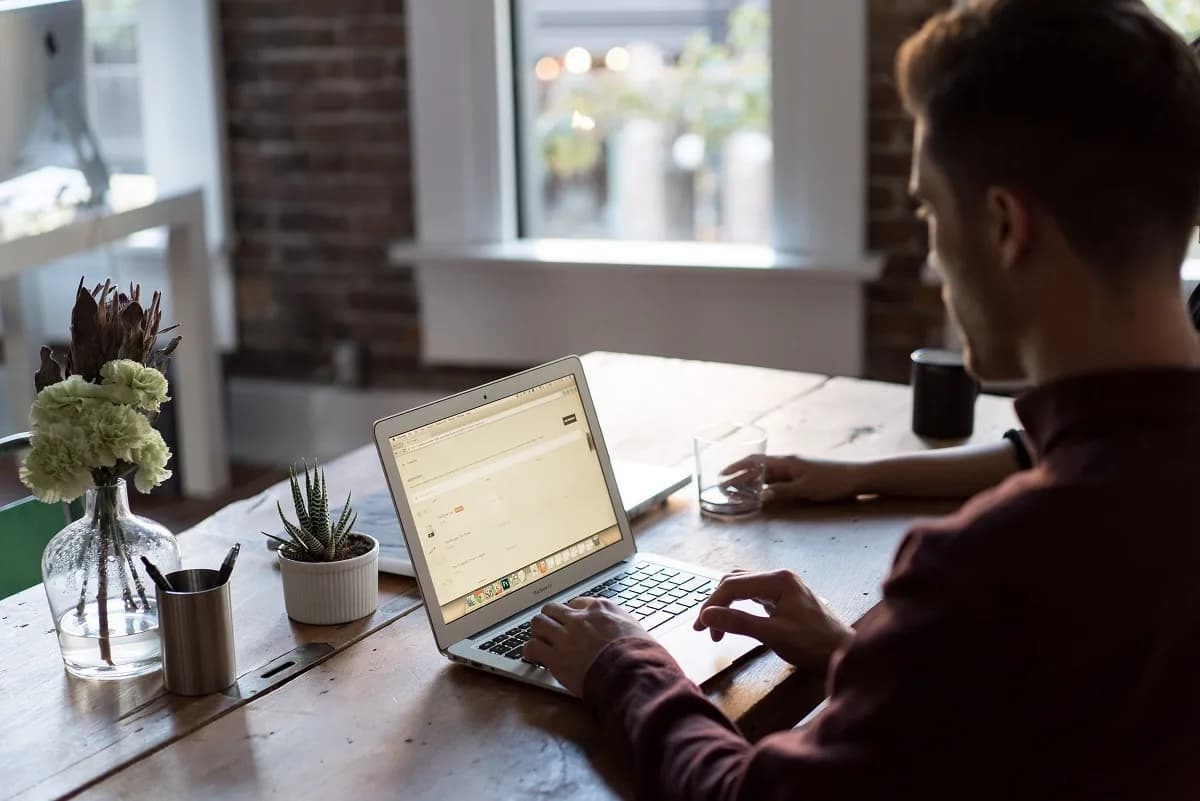 A man works at a laptop on a wooden table beside flowers in a bright room