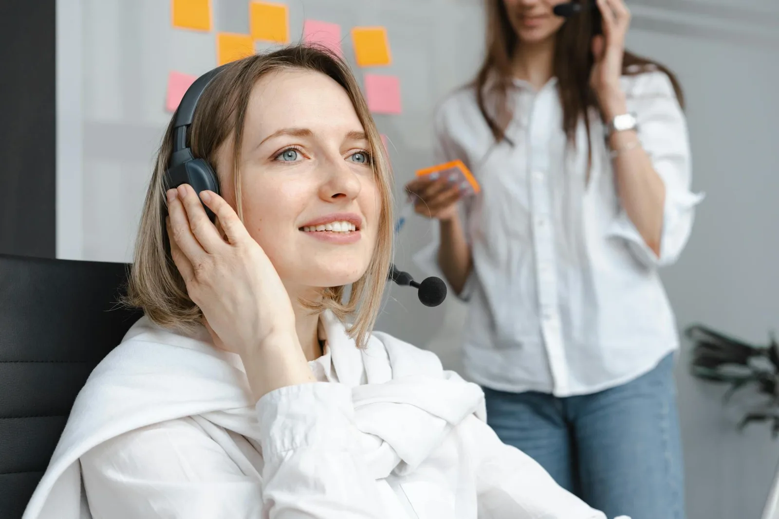 Two call-centre workers in headsets in a bright office with sticky notes behind them