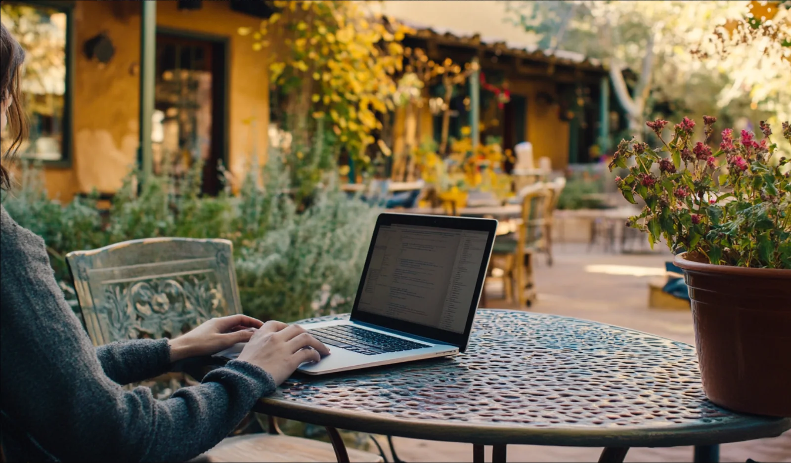 A person works on a laptop at an outdoor table beside an adobe building and plants